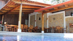 Un área de piscina con sillas y mesas de madera bajo una pérgola de madera.