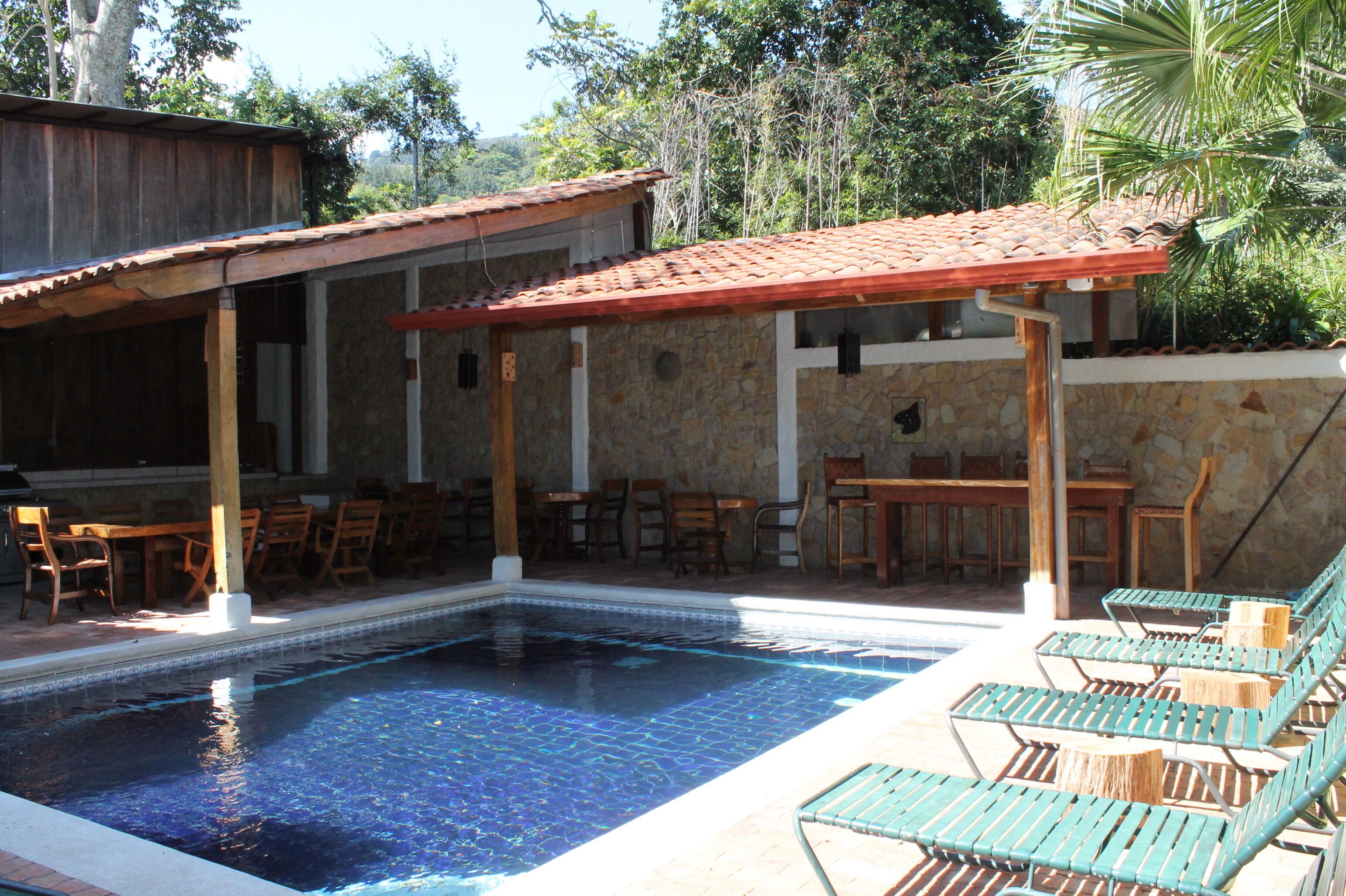 A pool area with lounge chairs, wooden tables, and a stone building with a tiled roof.