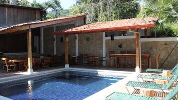 A pool area with lounge chairs, wooden tables, and a stone building with a tiled roof.