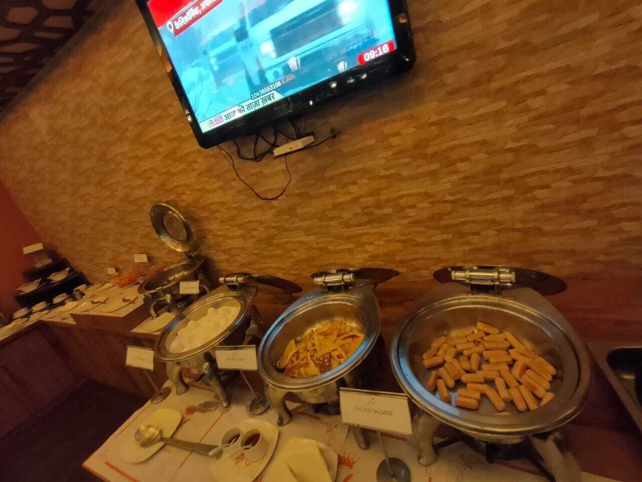 A buffet setup with labeled containers of food, a television displaying a news channel, and a wooden wall in the background.
