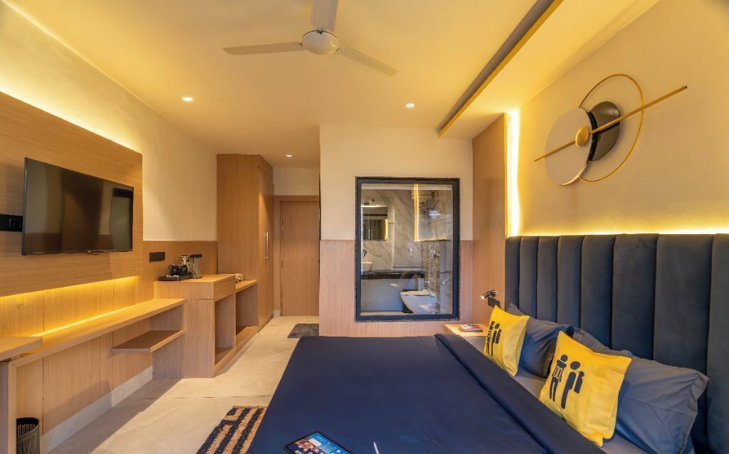 Un dormitorio moderno con una cama grande, un televisor en la pared, un escritorio y un baño visible a través de una puerta de cristal.