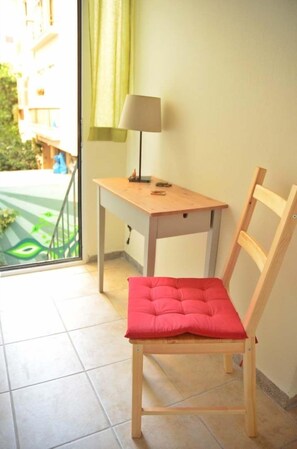 Une pièce comprenant une chaise en bois avec un coussin rouge, une petite table en bois avec une lampe, et une vue sur un espace extérieur verdoyant, visible à travers une fenêtre.