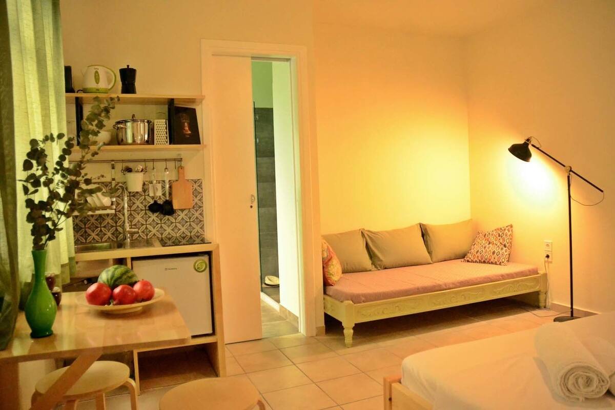 A compact room with a kitchenette, a sofa bed, a dining table with chairs, and a bedside lamp.
