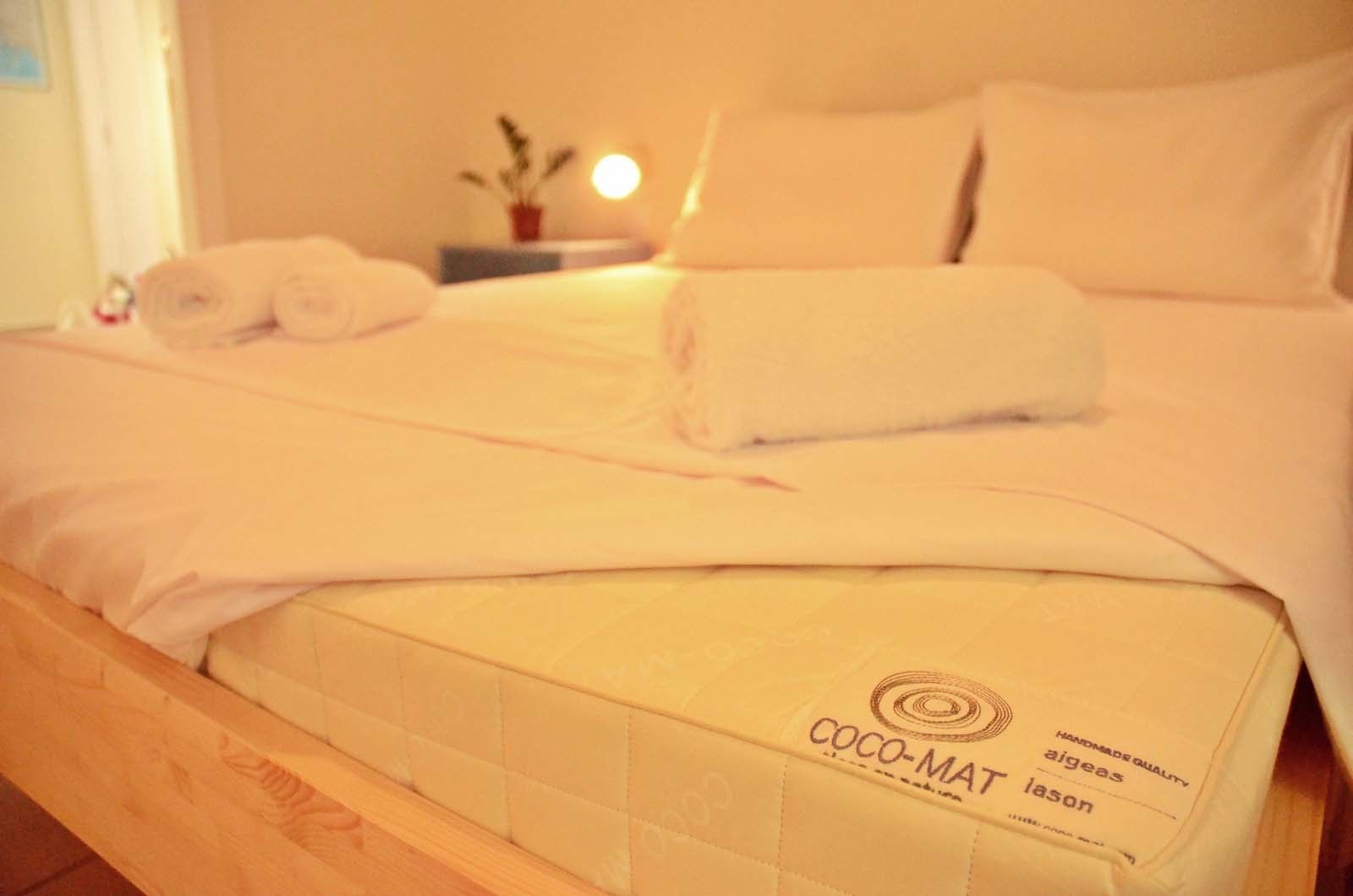 A neatly made bed with a white duvet and two rolled towels, a bedside table with a lamp, and a small potted plant.
