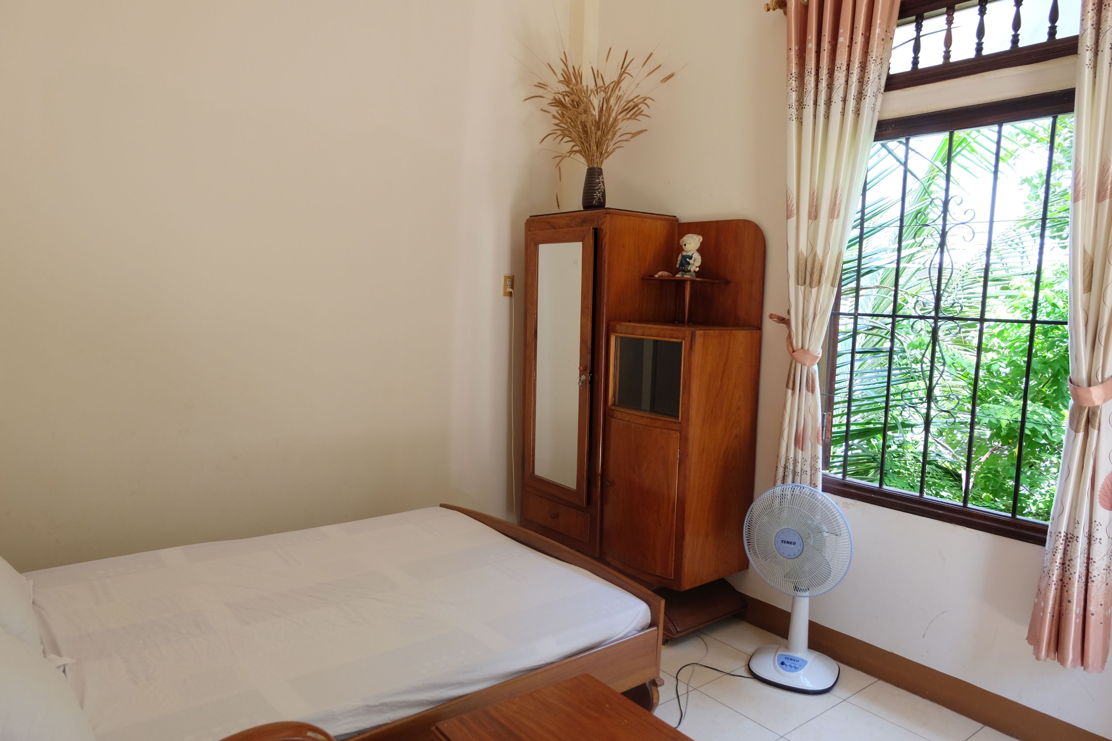 A bedroom with a bed, a wooden wardrobe, a fan, and a window with a view of greenery.
