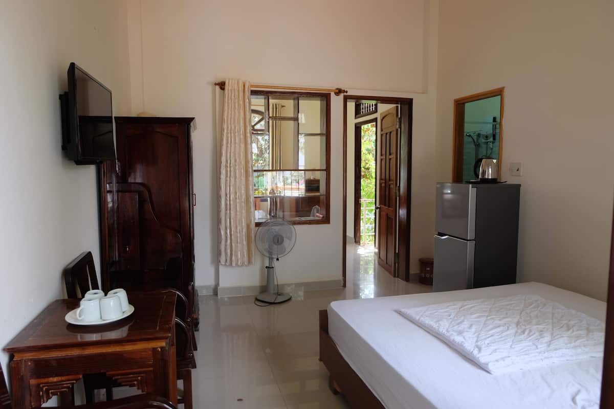 A hotel room with a bed, a desk with a chair, a television mounted on the wall, a fan, a refrigerator, and a window with curtains.