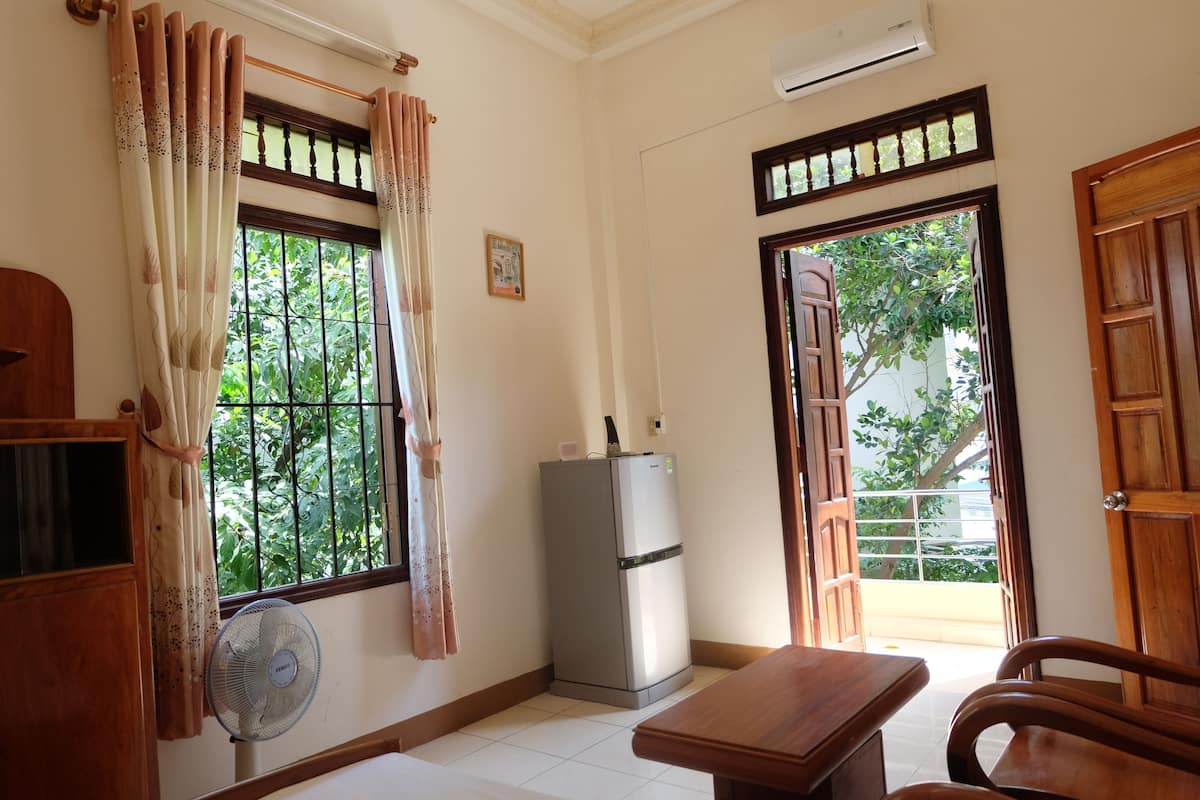 A room with a wooden table, a fan, a refrigerator, and a window with curtains.