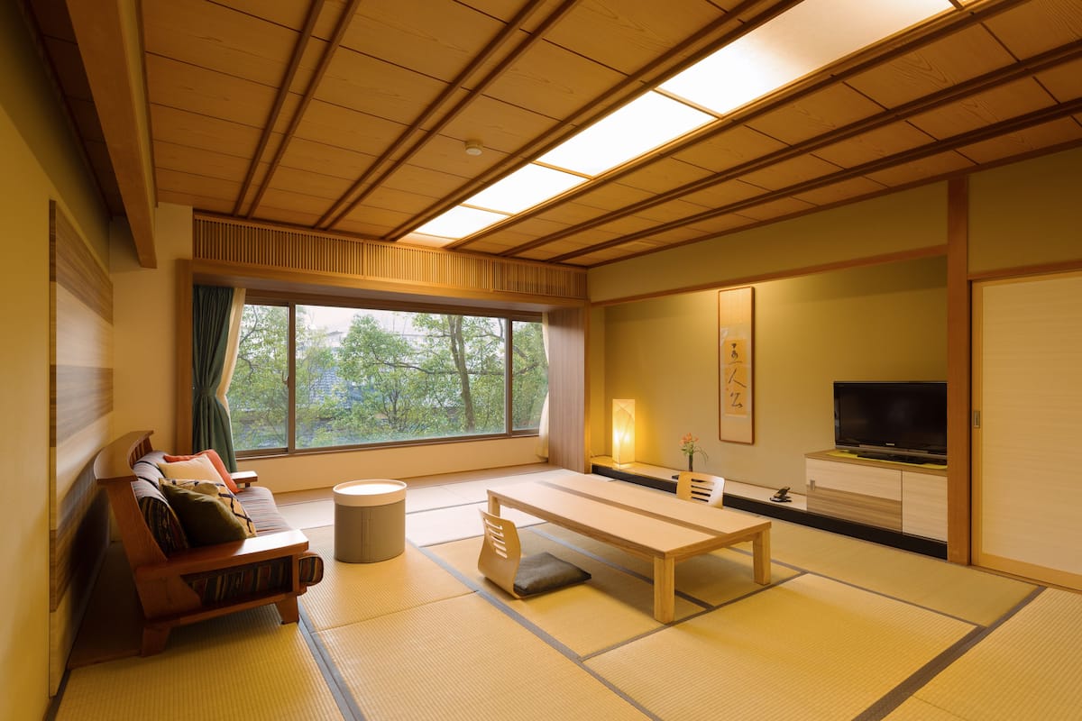 A traditional Japanese-style room with tatami flooring, a low wooden table, a sofa with cushions, a television, and a window with a view of trees.