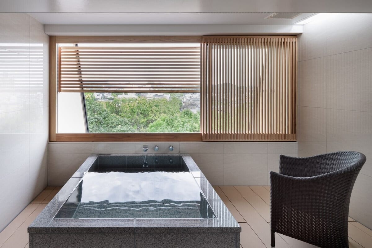 A modern bathroom with a glass-enclosed hot tub, a wooden window with blinds, and a wicker chair.