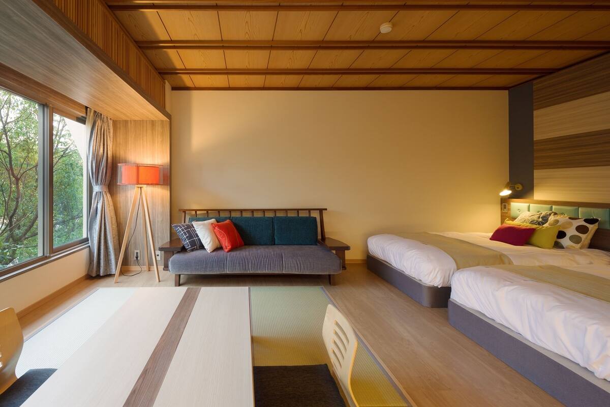 A modern hotel room with a sofa, two single beds, a desk, and a view of trees through the window.