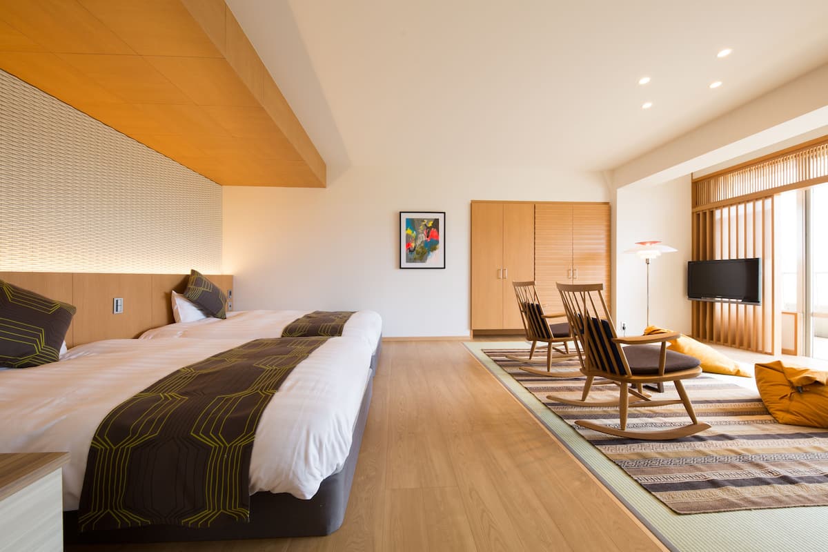 A modern hotel room with two beds, a wooden ceiling, a large window, a flat-screen TV, and a dining area with a table and chairs.