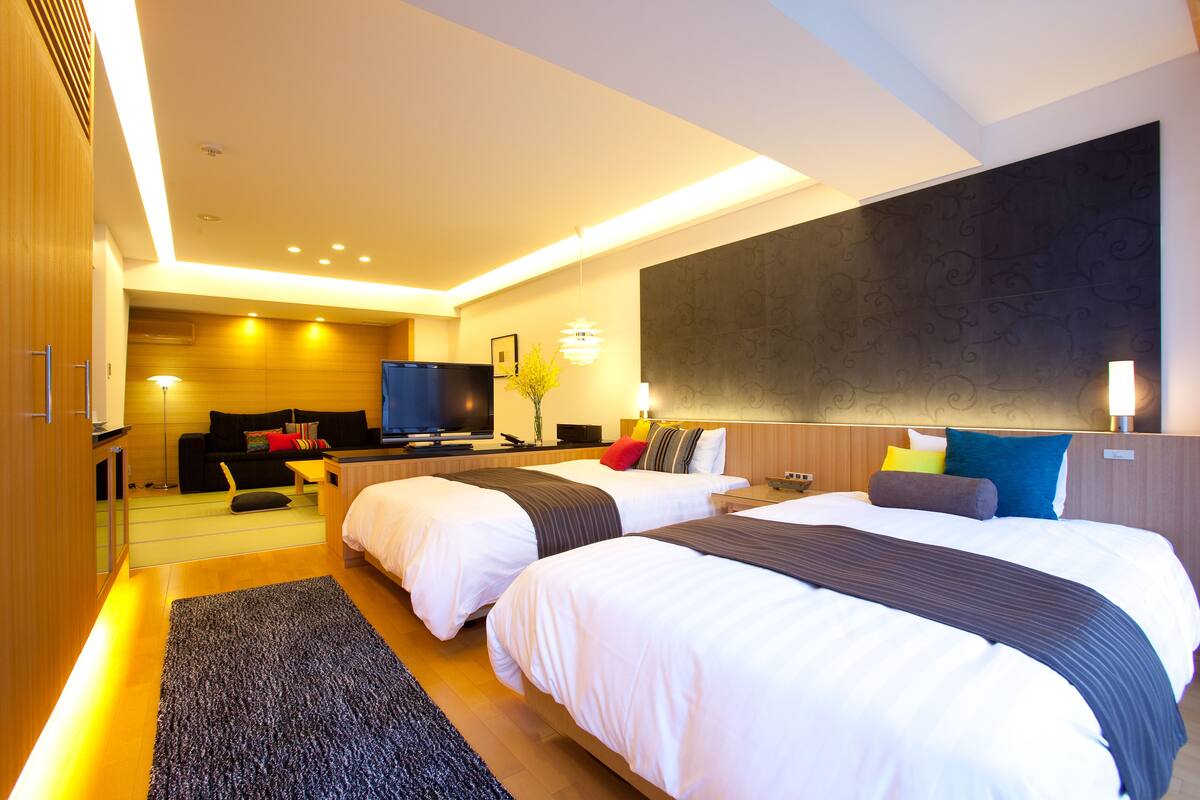 A modern hotel room with two beds, a TV, a sofa, and a large wall-mounted TV.