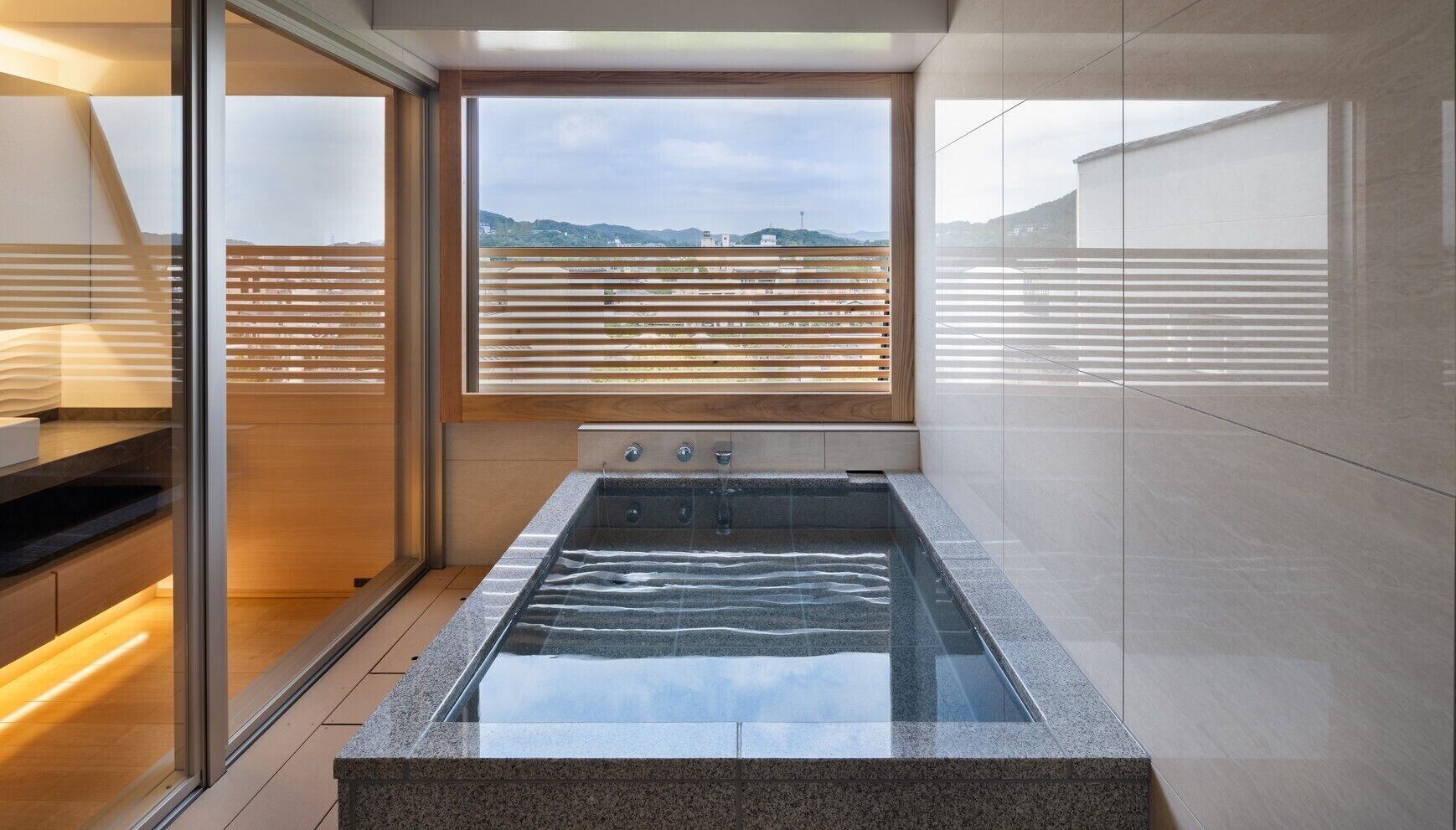 A modern bathroom with a glass-enclosed hot tub, wooden blinds, and a view of the cityscape.