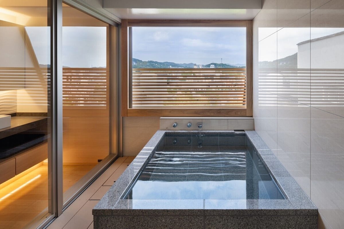 A modern bathroom with a glass-enclosed hot tub, wooden blinds, and a view of the cityscape.