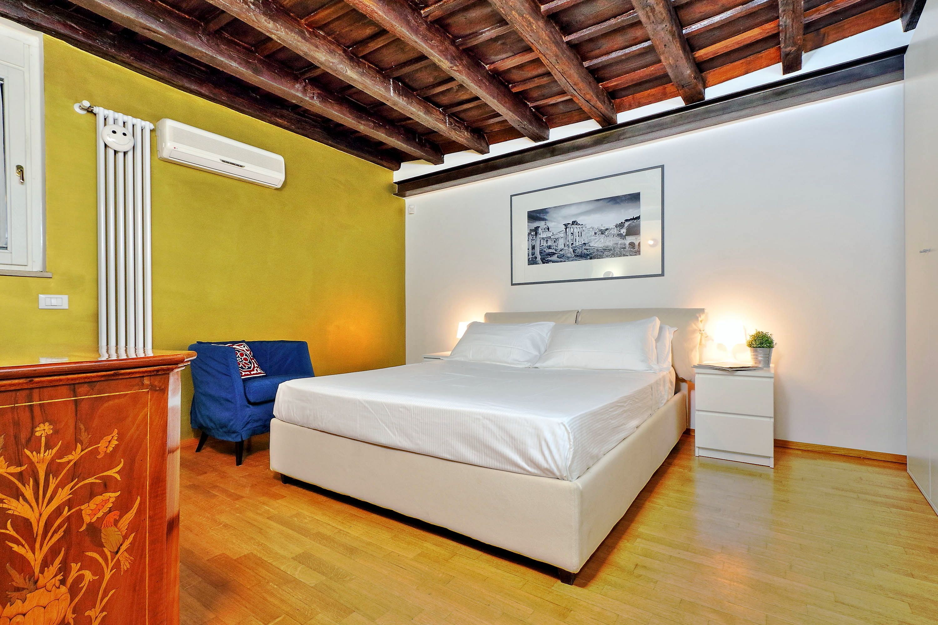 A bedroom with a wooden floor, a bed, a nightstand, a radiator, and a framed picture on the wall.