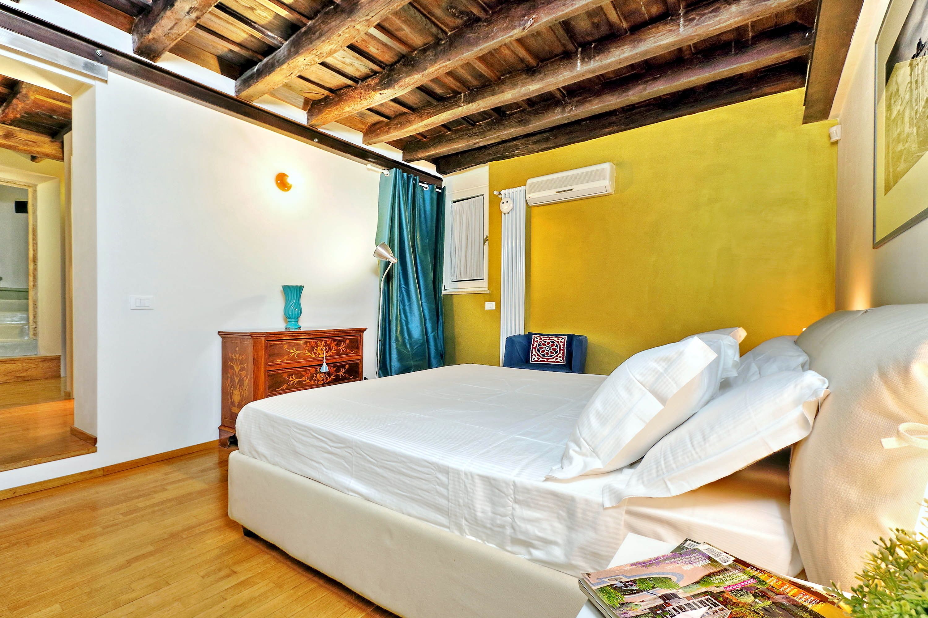Un dormitorio con techo de madera, una cama con ropa de cama blanca, una mesita de noche de madera, una silla azul y una pared amarilla.