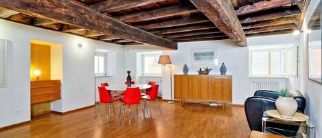 A spacious room with wooden flooring, a dining area with red chairs, a wooden cabinet, and a black leather chair.