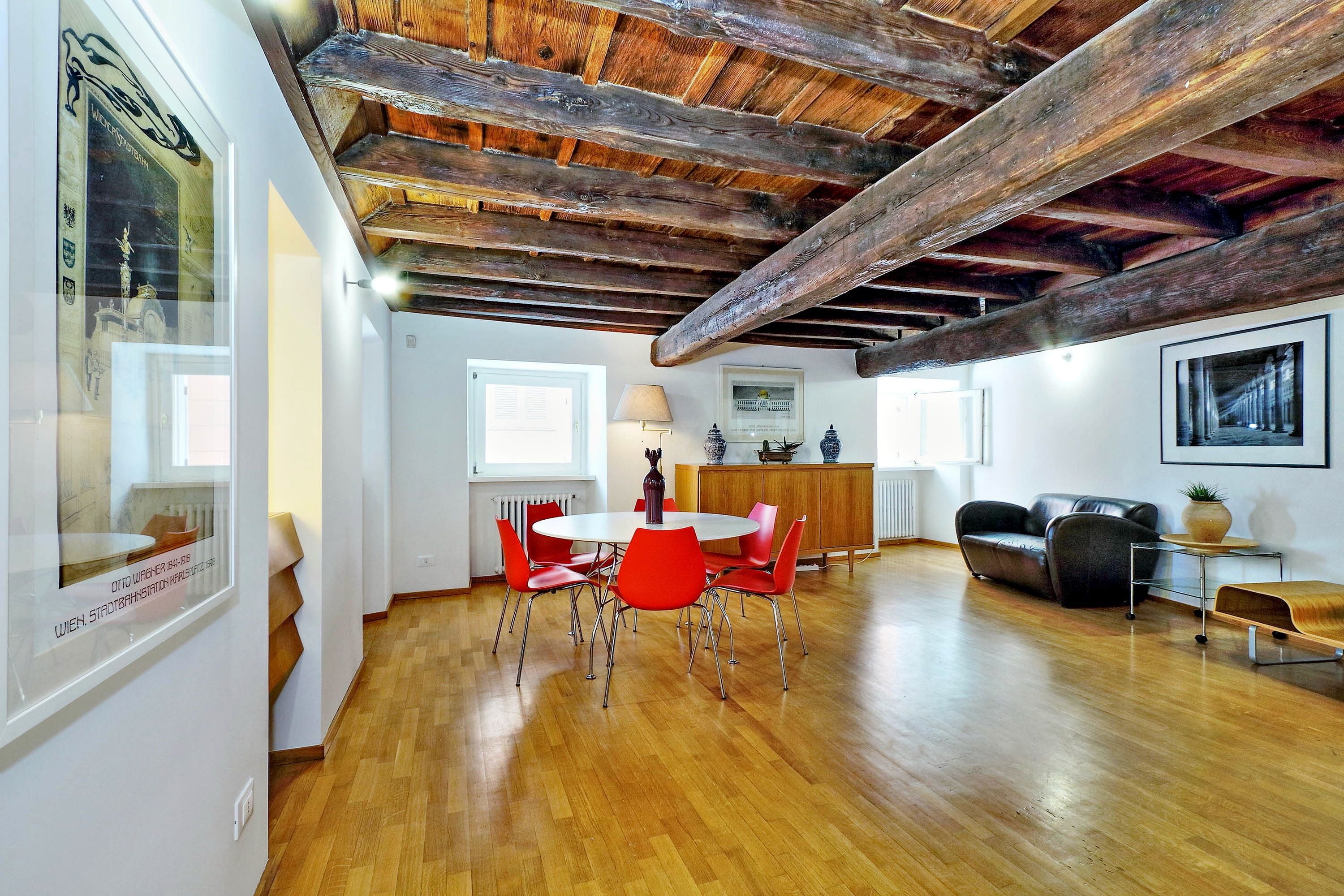 A spacious room with a wooden ceiling, a dining table with red chairs, a sofa, and a cabinet.