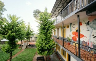 Un edificio con balcón, un mural con aves y flores, y un jardín con árboles y un sendero.
