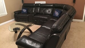 En sort lædersofa med to blå puder, et glasbord og fjernbetjeninger på det.