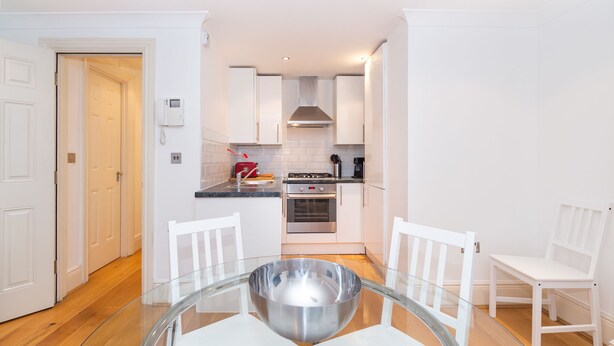 A compact kitchen with white cabinets, a gas stove, an oven, and a coffee maker. There is a glass table with chairs in the foreground. - PML Apartments Park Road (London)