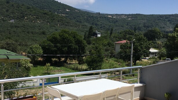 Una terraza con mesa y sillas blancas, con vista a un paisaje montañoso con vegetación y edificios.