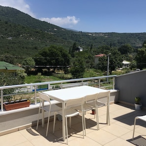 Une terrasse avec une table et des chaises blanches, donnant sur un paysage montagneux verdoyant, parsemé de bâtiments.