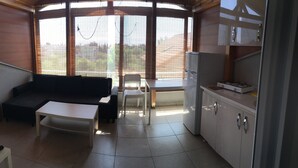Un espacio habitable compacto con sofá, una mesita, refrigerador y ventana con vistas a un barrio residencial.