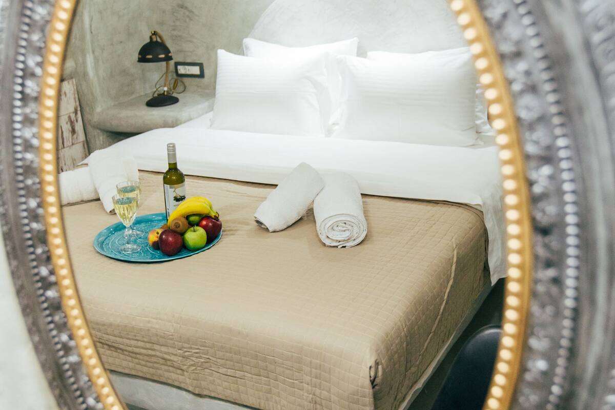 Een netjes opgemaakt bed met witte lakens, een opgerolde handdoek en een fruitschaal met een fles wijn.