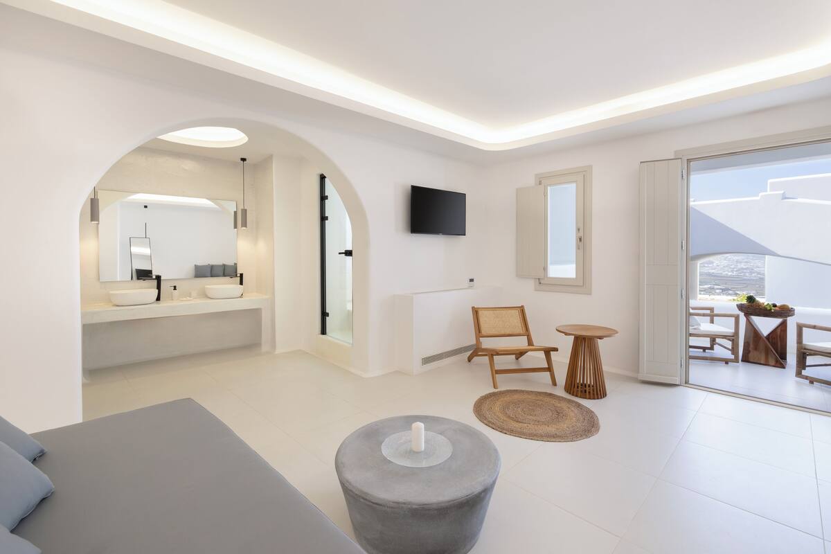 Een moderne hotelkamer met een ruime boogvormige deuropening naar een badkamer, een flatscreen televisie aan de muur en een balkon met zitplaatsen buiten.