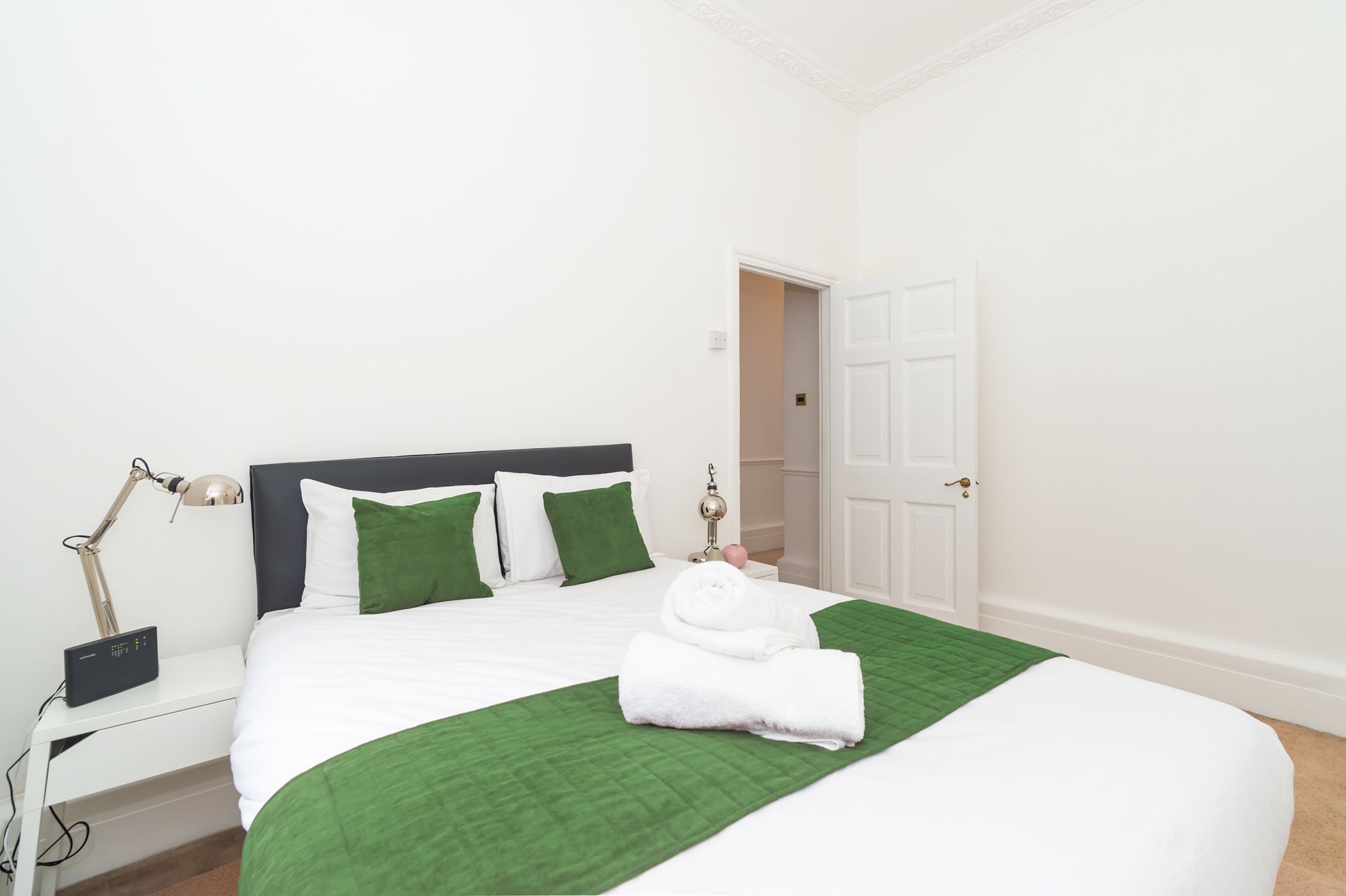 A neatly made bed with white bedding and green pillows, a bedside lamp, and a white door in the background.