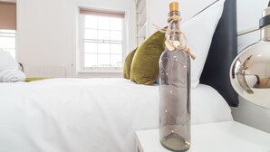Una botella de vidrio transparente con tapón de corcho y cuerda, sobre una mesita de noche blanca en una habitación de hotel.