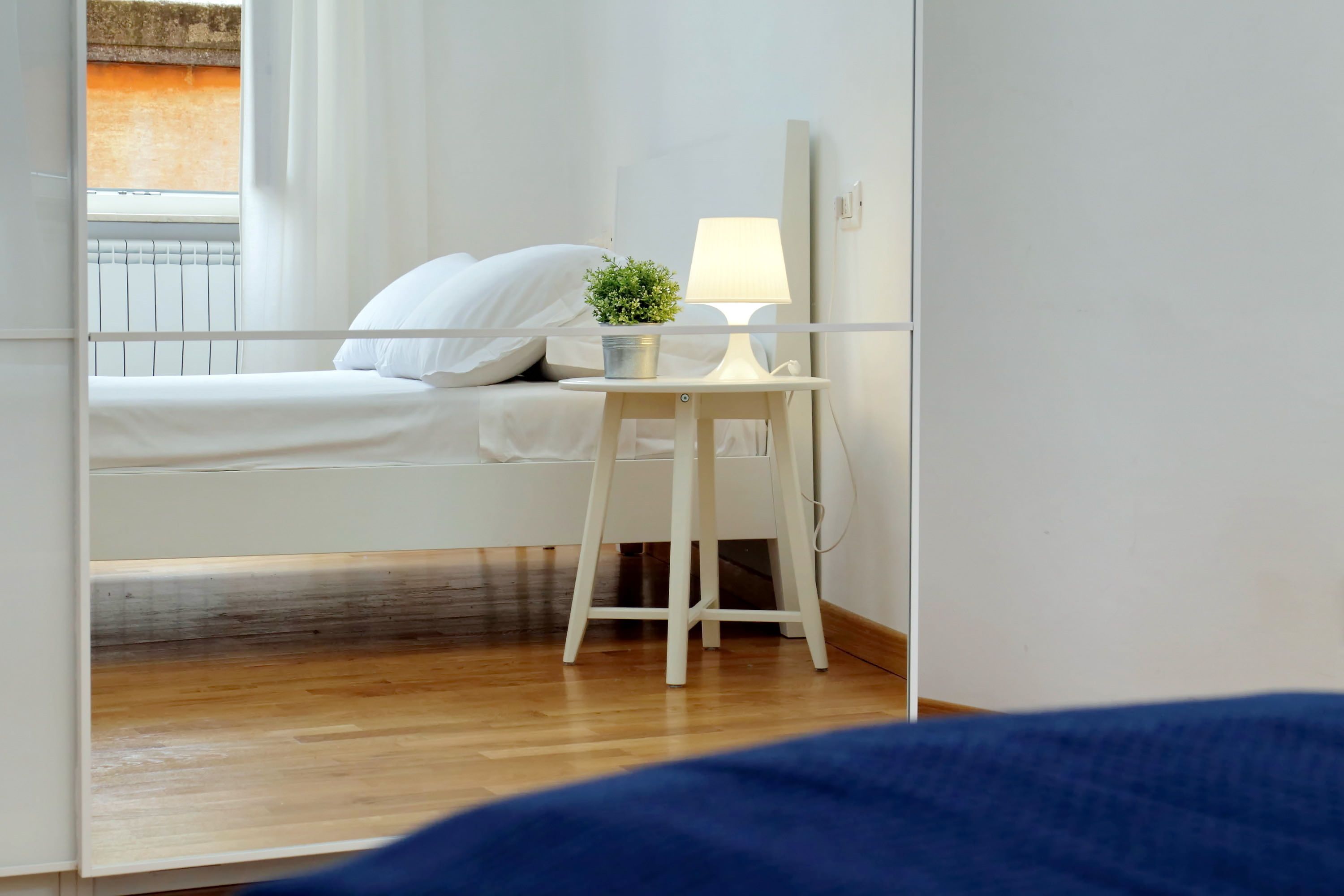 Un dormitorio con cama, una mesita de noche con lámpara, una pequeña planta en maceta y una ventana con cortinas.