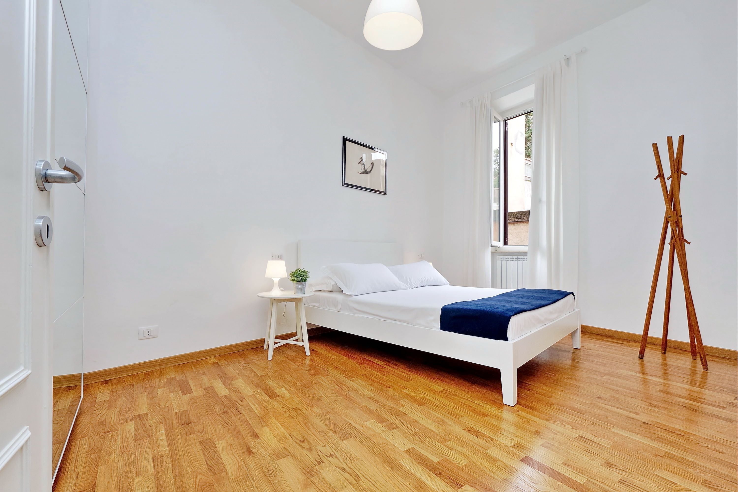 Une chambre moderne avec un lit blanc, un plancher en bois, une petite table de chevet avec une lampe, un tableau encadré au mur et une fenêtre avec des rideaux.