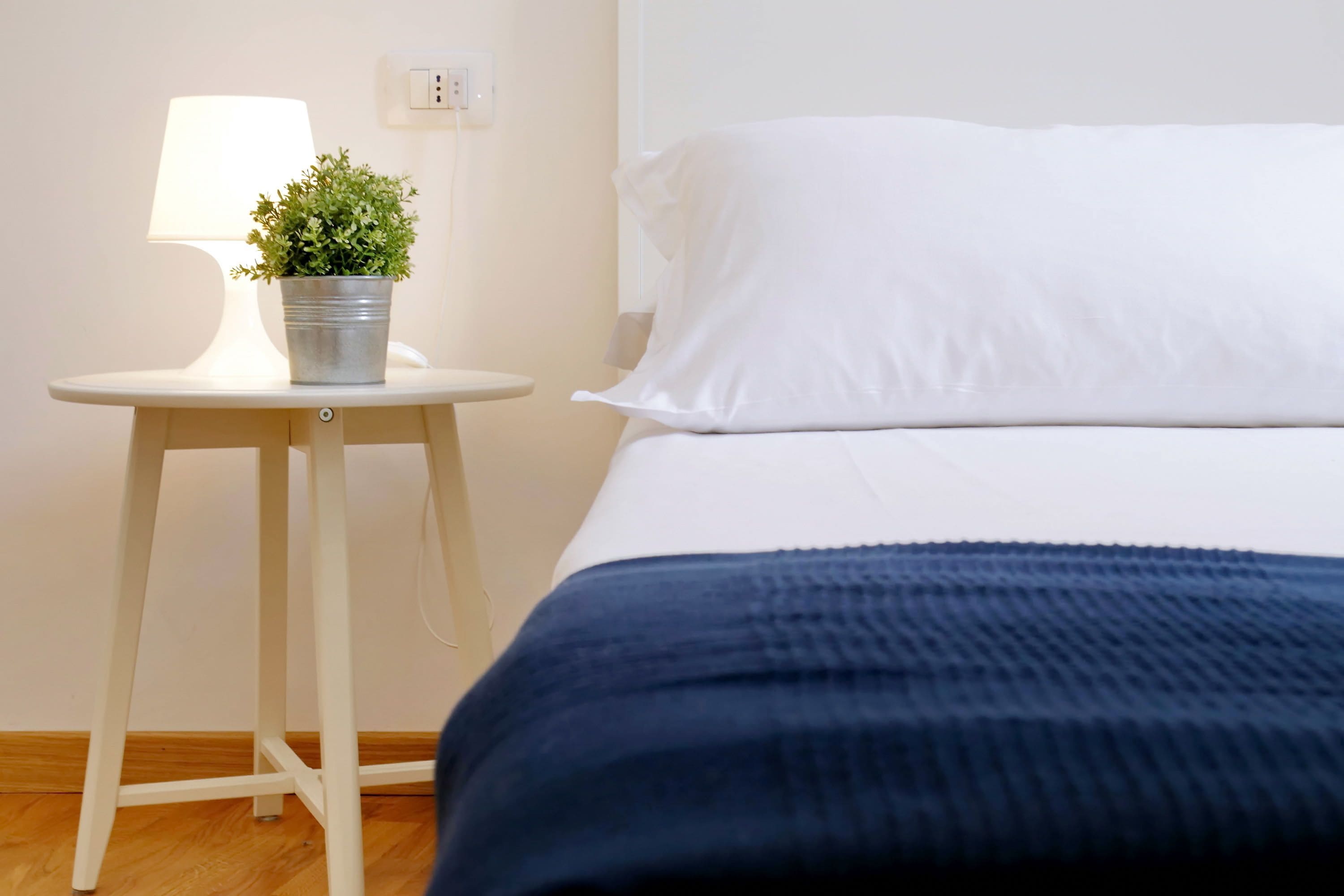 Een net opgemaakt bed met witte lakens en een donkerblauwe deken, een klein rond tafeltje met een lamp en een bloempot, en een houten vloer.