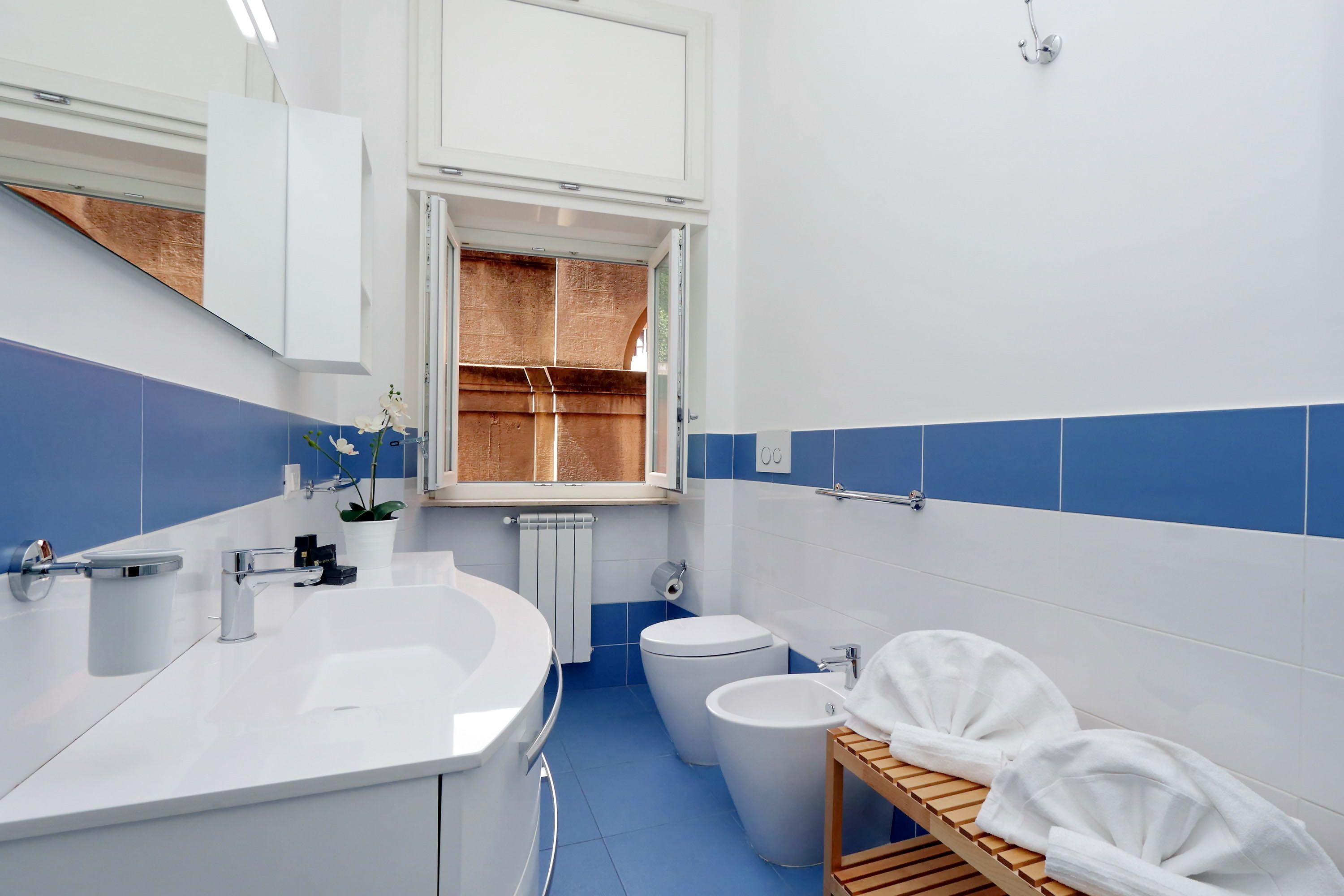 A modern bathroom with a white sink, a toilet, and a bidet. There is a wooden shelf with towels and a potted plant.