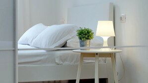 Een netjes opgemaakt bed met witte beddengoed en kussens, een kleine potplant op een nachtkastje, en een bedlampje.