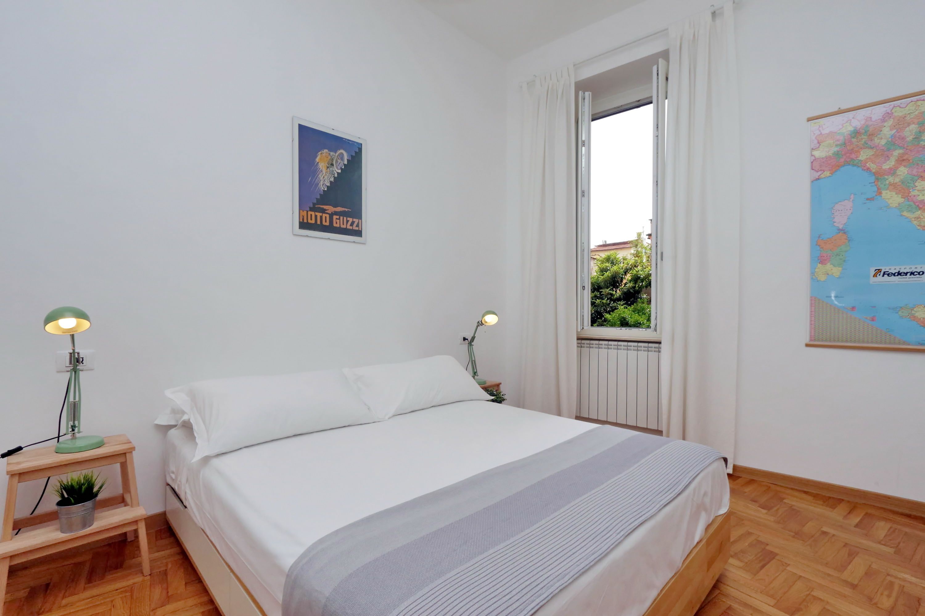 Un dormitorio con una cama, dos mesitas de noche con lámparas, un mapa en la pared y una ventana con cortinas.