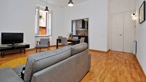 Een moderne woonkamer met een grijze bank, een flatscreen televisie en een eethoek met stoelen en een tafel.