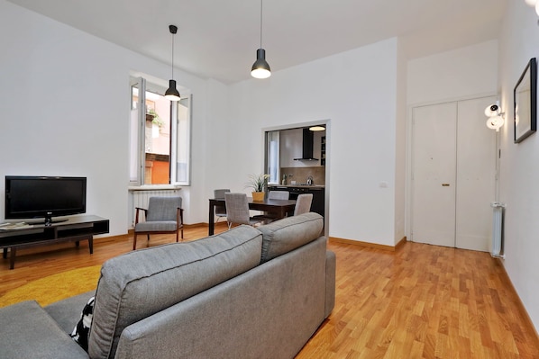A modern living room with a grey sofa, a flat-screen TV, and a dining area with chairs and a table.