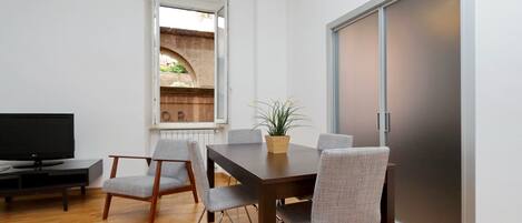A modern living room with a wooden floor, a dining table with four chairs, a television, and a large sliding door.