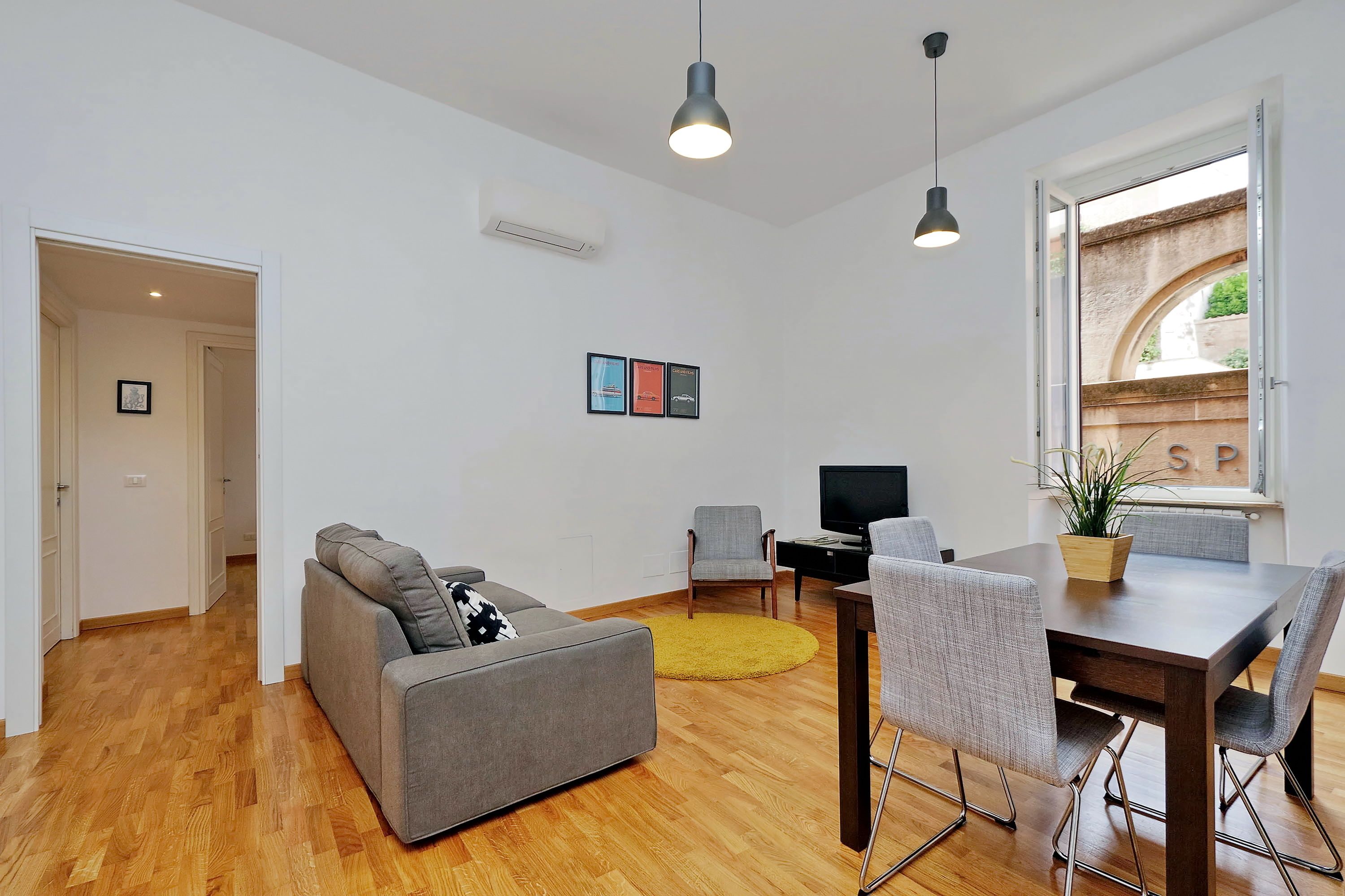 A modern living room with a grey sofa, a dining table with chairs, a TV, and a window with a view of a brick wall and greenery.