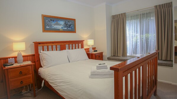 Habitación de hotel con una cama de madera, dos mesitas de noche con lámparas, un cuadro en la pared y una ventana con cortinas.