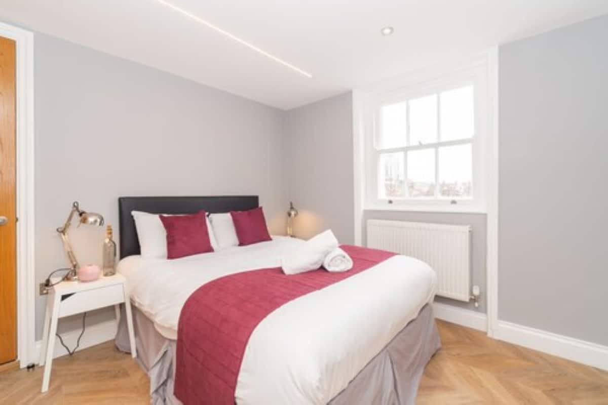 A neatly made bed with a red and white bedspread, a bedside lamp, and a window with a view of the city.
