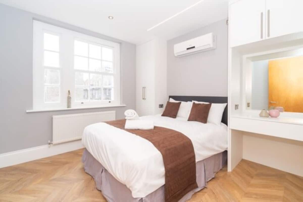 A modern bedroom with a large bed, a window with a view, an air conditioning unit, and a white dresser.