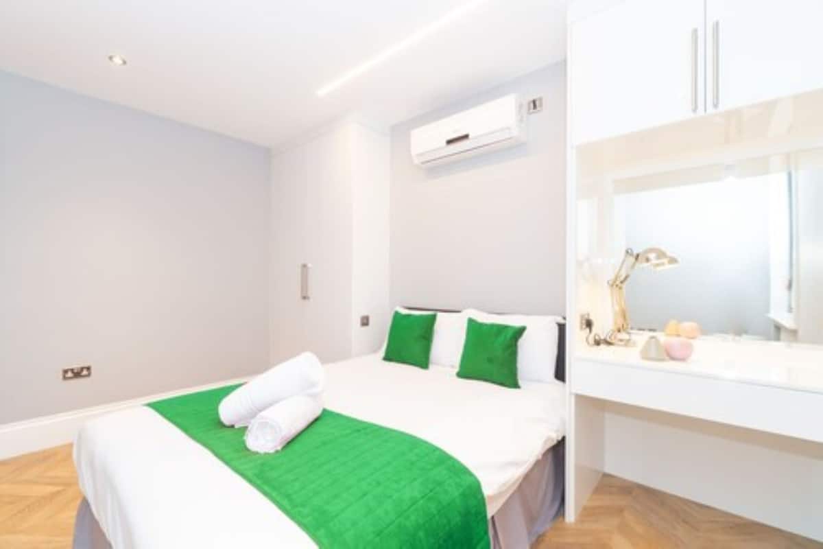 A modern hotel room with a bed, green pillows, a white headboard, a bedside table with a lamp, and a window.