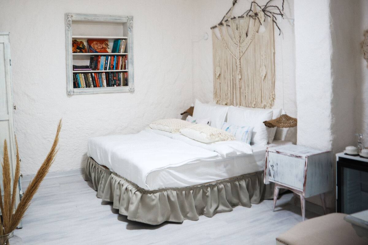 A bedroom with a bed, a bedside table, a bookshelf, and a macrame wall hanging.