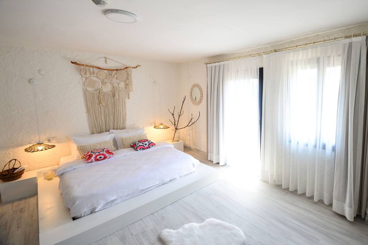 A modern bedroom with a large bed, bedside lamps, a decorative wall hanging, and sheer curtains.
