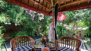 Un banc en bois avec des coussins à carreaux, une table garnie d’assiettes, et un parasol rouge.