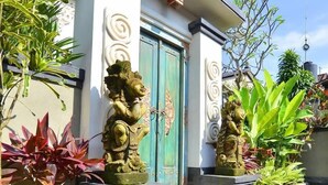 L’entrée d’un bâtiment, avec une porte bleue, flanquée de deux statues en pierre et entourée de différentes plantes.